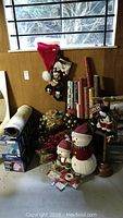 Multiple boxes of Noma 70-LED Christmas light strings stacked on floor, rolls of wrapping paper standing upright, plush snowman decorations, wreath, stockings with snowmen, and Santa hat on a wooden floor near window