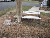 White metal bicycle planter with three wire baskets and a weathered wooden garden bench with white paint and metal supports on dry grass near a tree.