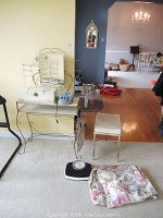 Full view of the vintage vanity with mirror and metal shelving, vanity bench with floral slipcover on cushion, mechanical scale on the carpet in front, and some other items around it.