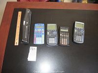 Full view of the four calculators, three hole punch, and ruler placed on a black surface