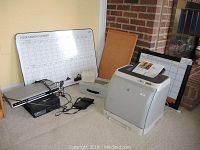 HP Color LaserJet 1600 printer with whiteboards and miscellaneous office supplies including power adapters and cables visible on carpeted floor near a brick wall.
