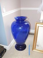 Full front view of the large cobalt blue glass floor vase placed on the carpeted corner of a room showing its size and shape.