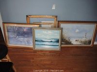 Five items displayed: three framed artworks and two empty frames leaning against a wall on a hardwood floor.