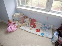 Two baby dolls with assorted doll clothing, accessories, and a small blanket displayed on carpet by window.