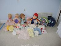 Five vintage dolls seated on carpet with assorted doll clothing and two vintage doll cases behind them