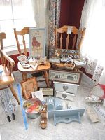 Full view of two wooden chairs with various country kitchen decorative items arranged on and in front of them. Items include framed prints, wooden signs, shelves, baskets, and small accessories.