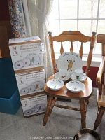 Three boxed 16-piece Merry Brite Christmas porcelain dinnerware sets stacked next to a wooden chair displaying some dishes of the set: one large dinner plate, two soup bowls, two cups, two saucers.