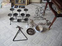 Photo showing a large black metal candle holder with nine platforms, a curved metal wine rack, a metal decorative dinner bell shaped like a triangle, two silver ornamental metal holders, a white and blue ceramic vase, and a decorative metal plate.