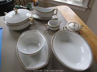 Complete set of white porcelain serving pieces with gold and black trim shown on wooden table under daylight. Includes bowls with lids, sugar bowl, creamer, gravy boat with underplate, butter dish with lid, and platters.