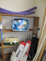 Full view of shelving unit with two golf bags filled with clubs, fishing tackle box, and pool noodles above, as well as gray tub with pool liner.