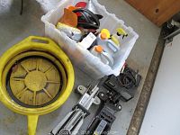 Assorted automobile items including bottles of fluids in plastic bin, yellow funnel, battery charger, and two floor jacks on floor.