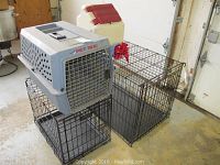 Two metal wire pet crates stacked; top crate has plastic tray; large plastic pet carrier positioned on top; white plastic food storage bin with red lid and scoop behind.