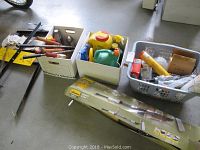 Overview of assorted hardware and garden items including hand tools, watering cans, and paint rollers arranged in boxes and on the floor.