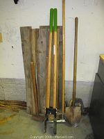Wooden fence posts leaning against wall alongside long-handled post hole digger, pole driver with green grips, and shovel.