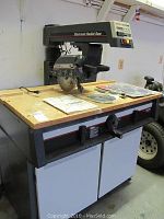 Radial saw mounted on cabinet with accessories laid on top, showing the saw, cabinet doors, and extra blades on the top surface.