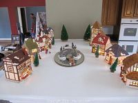 Wide shot of several ceramic houses with lights on, miniature pine trees, and the bridge with people figurines arranged on a table to show the lot contents.