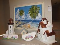Wide view of three decorative birdhouses with white bodies and brown roofs, floral painted smaller birdhouses with curved bases, and two palm tree beach coastal paintings framed on wall.