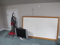 Photo showing large whiteboard with wooden frame, upright Eureka Electrolux vacuum cleaner, small portable heater, and 20-inch Sylvania monitor together in one view.