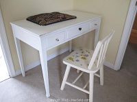 White wooden desk with three drawers and round knobs, visible chips and scratches on edges. Matching chair with floral painted seat and slatted back. Patterned cushion on desk.