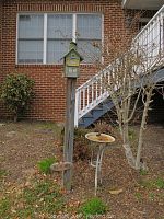 Wooden welcome sign mounted on a 65-inch tall wooden pole with a green birdhouse on top located in a garden bed near house stairs.