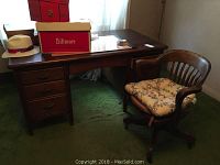 Wooden desk with three drawers on the left and accompanying wood office chair with floral cushion on green carpet.