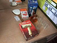 Overview of all items on table: boxed hair dryer, beard shaver, bottles of cologne, Band-Aid box, hand mirror, and four glass shot glasses.