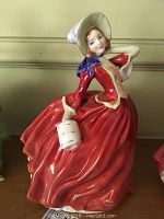 Front view showing the woman in a flowing red dress, bonnet hat, and holding a small white basket.