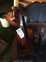 Full view of violin leaning on a blue upholstered chair, showing body, neck, scroll, with sticker labeled 55 visible