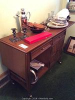 Wood server from front angle, showing foldable leaf on left and items on top including candlesticks, cocktail shaker, wooden bowls, metal tray and plates.