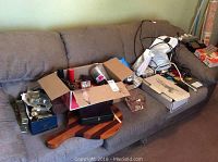 Wide photo showing lots of assorted household items on couch including boxes, cutting board, cables, flashlights, and other small items.