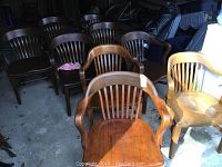 Nine wooden chairs arranged in a garage, front chairs with curved arms and slatted backs, various stain tones.