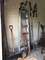 Photo showing the collection of garden and household tools including a 6-foot aluminum ladder, various shovels, pruning shears, rake, broom, brush, and hedge trimmer hanging on the wall or leaning against it.