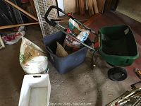 Overview photo showing the spreader with its green hopper and black frame alongside several bags of lawn care products placed in a gray plastic bin and on the ground, including Scotts Crabgrass Doctor and partially opened bags.