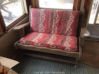 Front view of vintage painted pine love seat with red patterned cushions, showing wood frame and armrests.