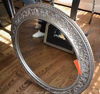 Photo of an ornate round mirror with an embossed floral and scroll pattern silver metal frame resting against a metal table leg on a wood floor.
