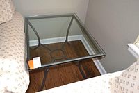 Square metal framed end table with glass top positioned between two chairs on wooden flooring.