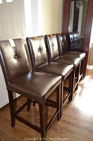 Four dark brown leather-like upholstered bar stools with wooden legs, arranged in a line on wooden floor. Each stool has a square padded seat and a padded backrest with a single button tuft centered.