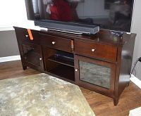 Wooden entertainment center TV console with TV and soundbar on top, two drawers, two glass door cabinets, and central open shelves visible front view.