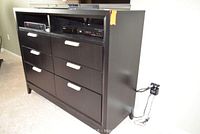 Front-right angled view showing six drawers, open shelves holding electronic devices, dark finish, silver handles, TV on top.
