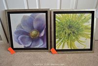 Two framed floral art pieces leaning against a door, showing a purple flower and a green flower with detailed petals, each marked with orange auction tags