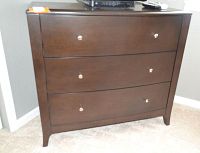 Front view of three-drawer wooden dresser with silver knobs placed against a grey wall on carpeted floor.