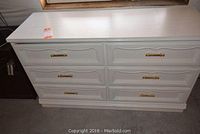 Front view of the white 6-drawer dresser showing ornate drawer faces and gold-tone handles.