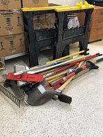 Photo showing pair of black and yellow plastic sawhorses and multiple yard tools including rakes, shovels, and post hole digger on floor.