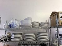 Three shelves of stacked white plates and bowls, some pasta bowls, displayed on a metal rack.