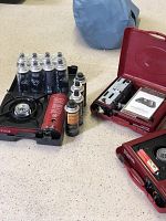 Three red and black portable butane grills on floor with 12 cans of fuel
