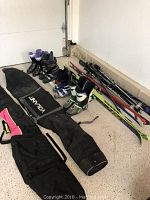 Photo showing three pairs of skis with bindings, four pairs of ski boots, poles, and ski bags arranged on the garage floor.