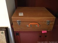 Closed view of wooden case with orange handle and metal latches showing minor hinge issues.