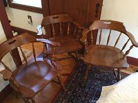 Three wood-tone dining chairs with curved armrests and spindle backs in a warm medium brown finish, placed on a wooden floor near a closed door and window. Shows general style and wood condition.