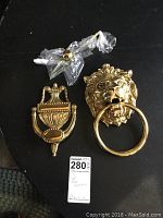 Two brass-colored door knockers including lion head and urn shapes, with included attachments on a black background.