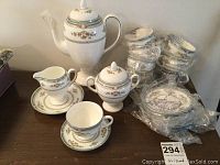 Complete tea service set arranged on a surface showing teapot, creamer jug, sugar bowl, and several stacked tea cups with saucers wrapped in plastic.